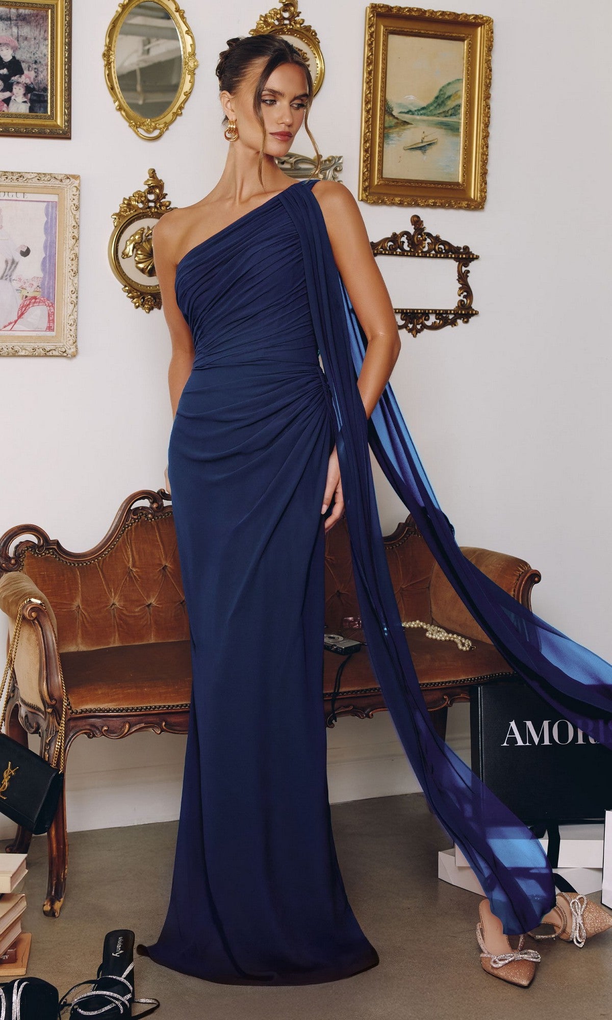 A woman wears the navy Amoris T730 long prom dress with a ruched bodice, standing in a vintage-style room featuring framed art, a wooden bench, and shopping bags on the floor.